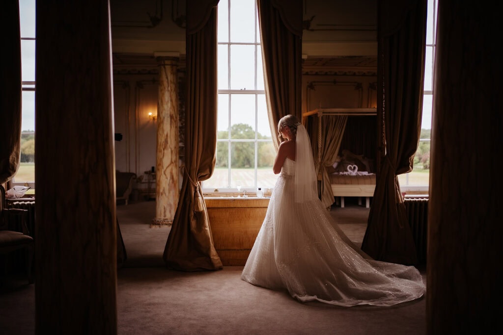 Bride looking out the window in a period bedroom at Gosfield Hall wedding venue — documentary bridal preparation photography by Lily & White