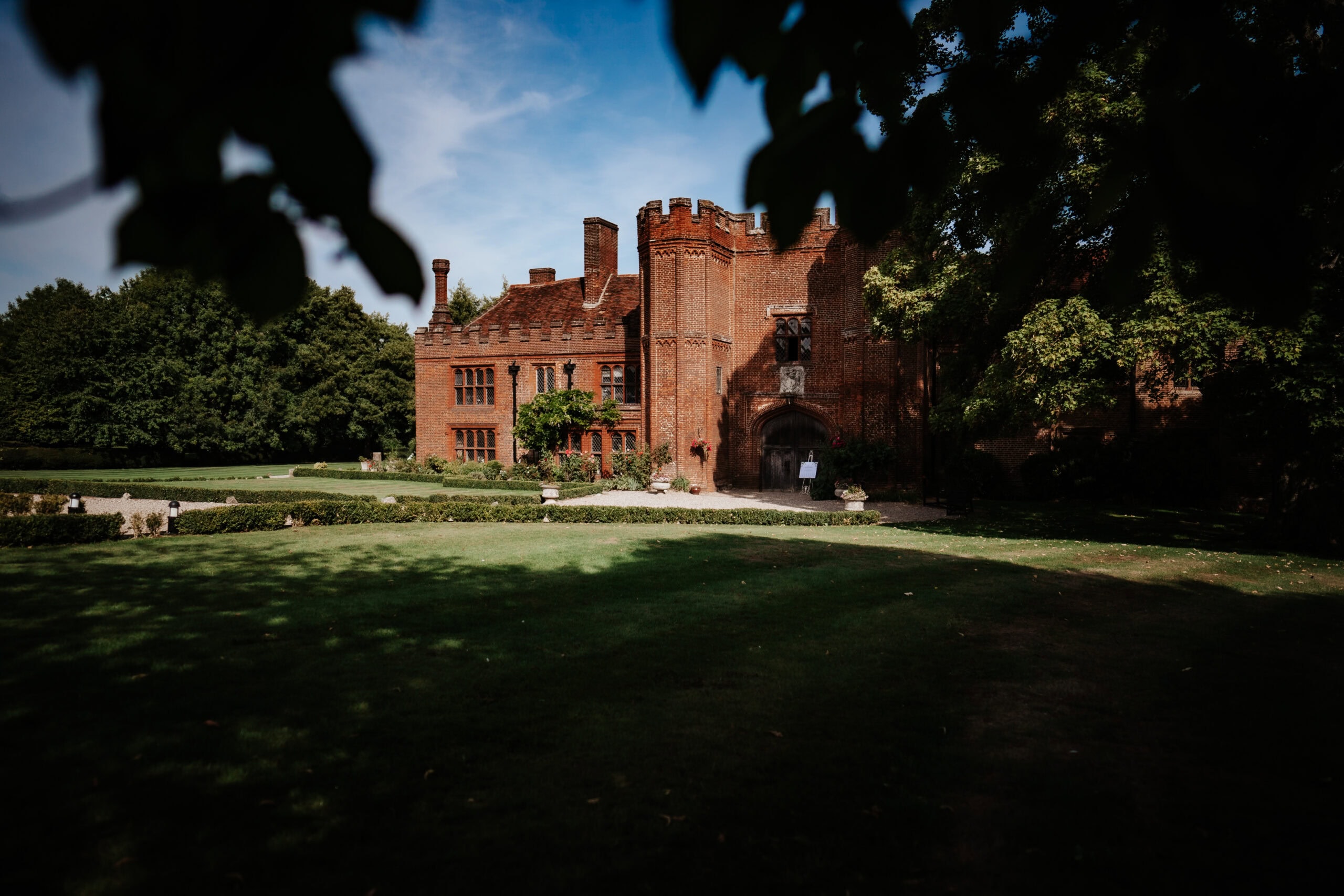 The Tudor gatehouse and Great Tower of Leez Priory wedding venue in Little Leighs, Essex, framed through overhanging tree branches on a summer day. The Grade I listed red brick manor house sits behind manicured lawns and topiary hedging. Photographed by Tel, Lily & White Photography, Leez Priory wedding photographer.