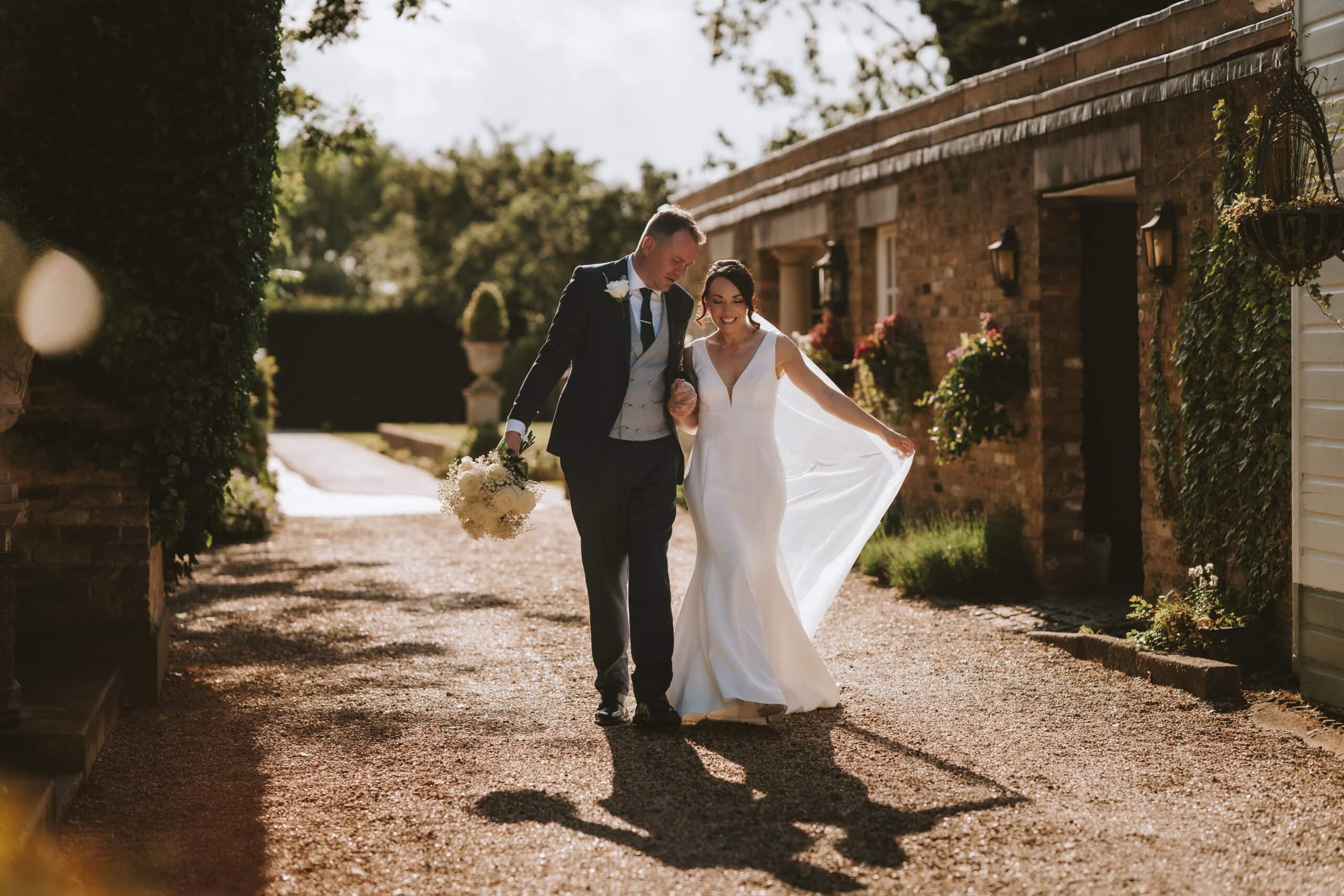 Bride and groom walking through the courtyard at Friern Manor country hotel in Brentwood with golden afternoon light – natural wedding photography by Lily & White