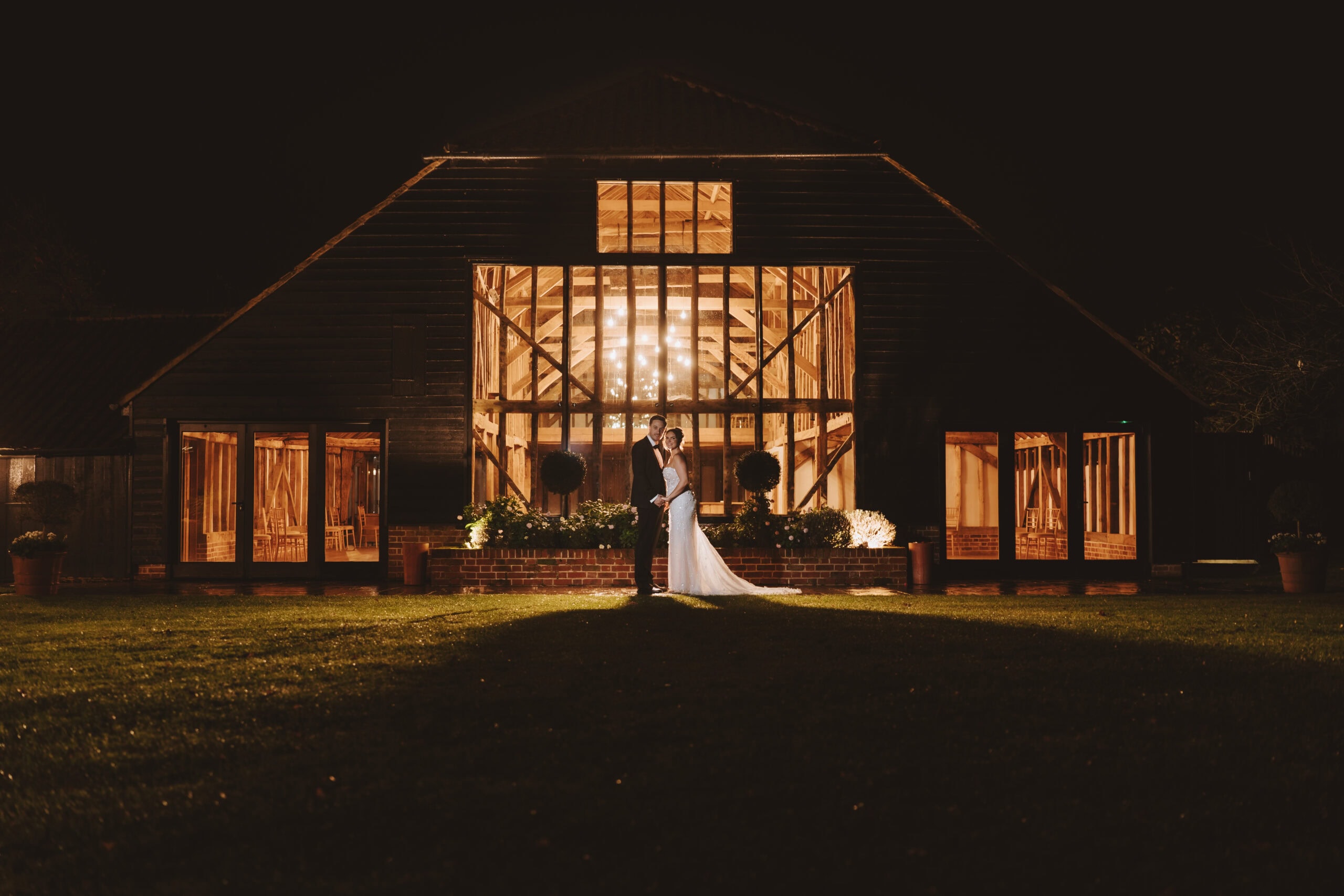 Abbey and Charlie standing together outside the illuminated Essex Barn at Blake Hall at night, warm light glowing through the full-height glazed gable behind them – wedding photography by Tel, Lily & White
