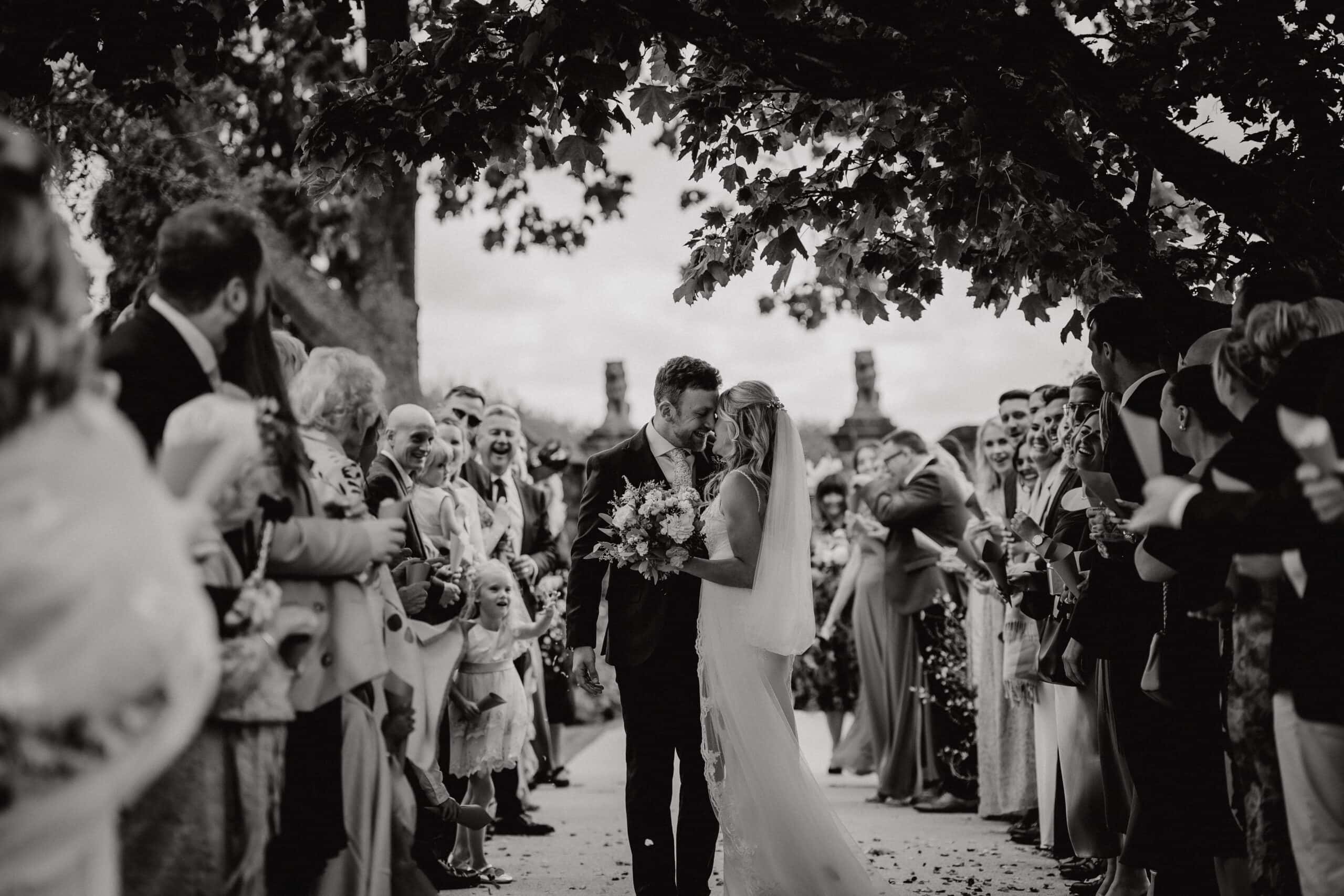 A black and white photograph of a bride and groom sharing a kiss during their confetti walk along the Long Walk aisle at Gaynes Park wedding venue in Epping, Essex. Guests line both sides of the stone path in celebration, with the ancient brick gateposts and overhanging tree framing the moment above. Photographed by Tel, Lily & White Photography, Gaynes Park wedding photographer.