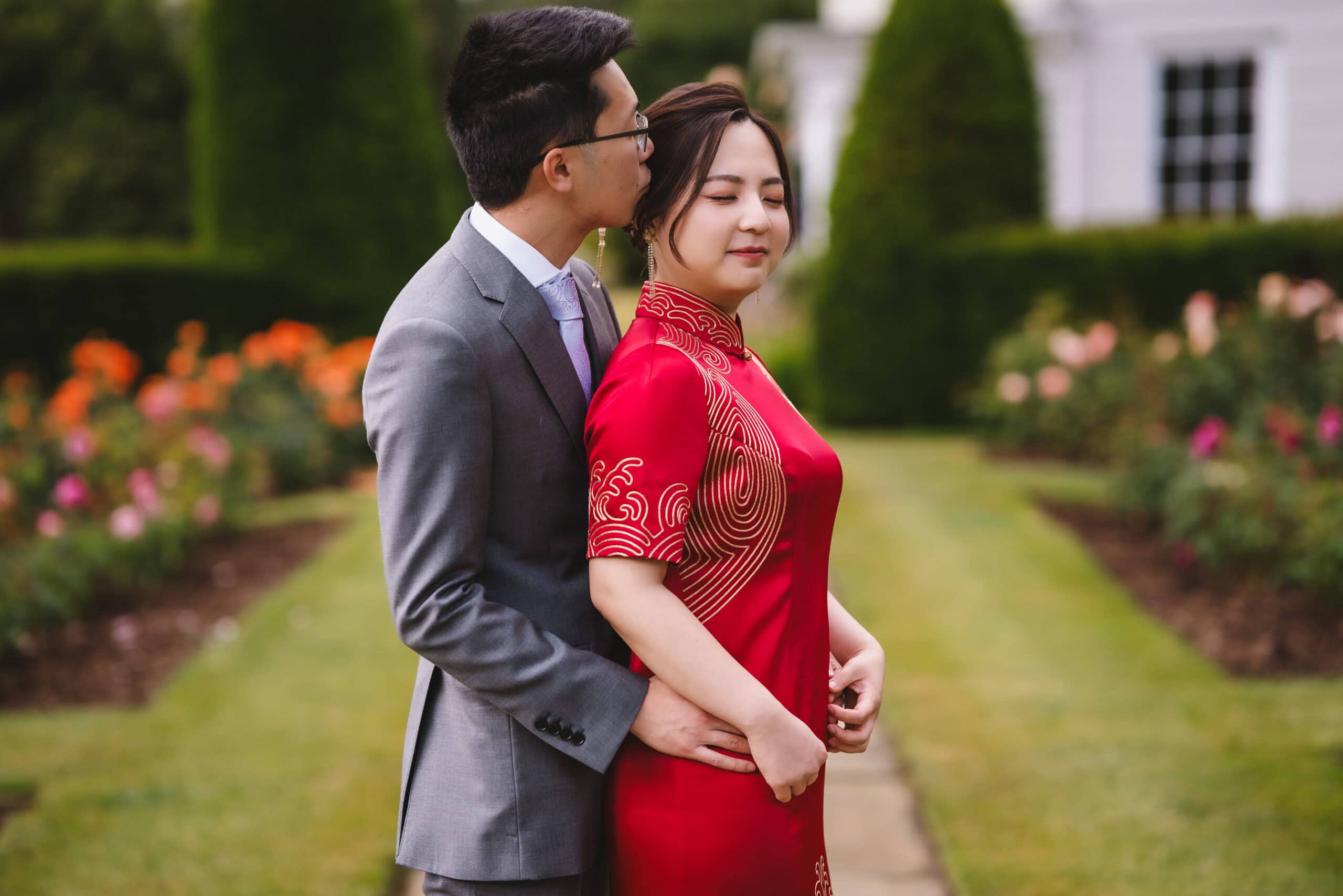 Yiran and Jeffrey in the rose garden at Blake Hall, Jeffrey kissing Yiran's cheek as she smiles in her red Chinese wedding dress – wedding photography by Tel, Lily & White