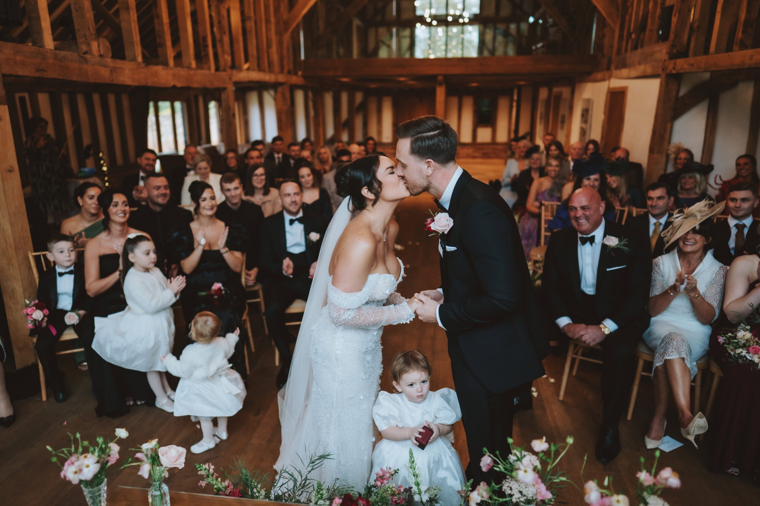 Abbey and Charlie sharing their first kiss during the wedding ceremony in the Hay Barn at Blake Hall, flower girls at their feet and guests applauding on both sides of the aisle – wedding photography by Tel, Lily & White