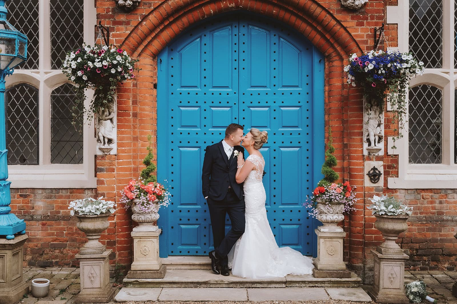 Bride and groom sharing a kiss in front of the iconic blue doors at Gosfield Hall, Essex, surrounded by stone urns filled with coral and pink flowers — wedding photography by Lily & White