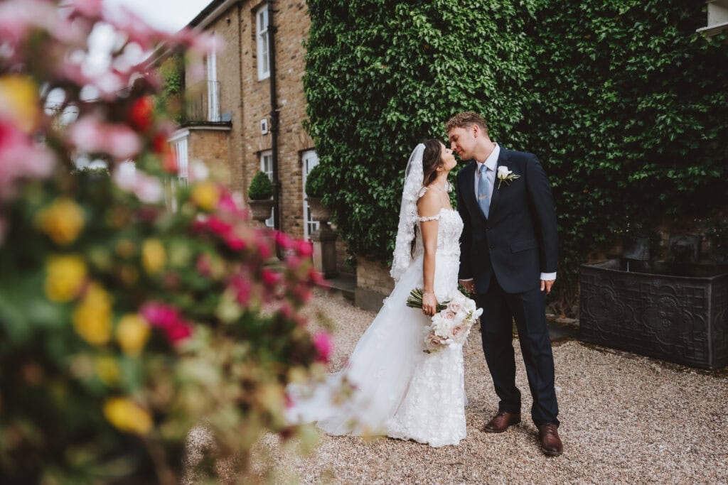 Bride and groom kissing in the courtyard at Friern Manor country hotel with ivy-covered building and flowers in foreground – romantic wedding photography by Lily & White