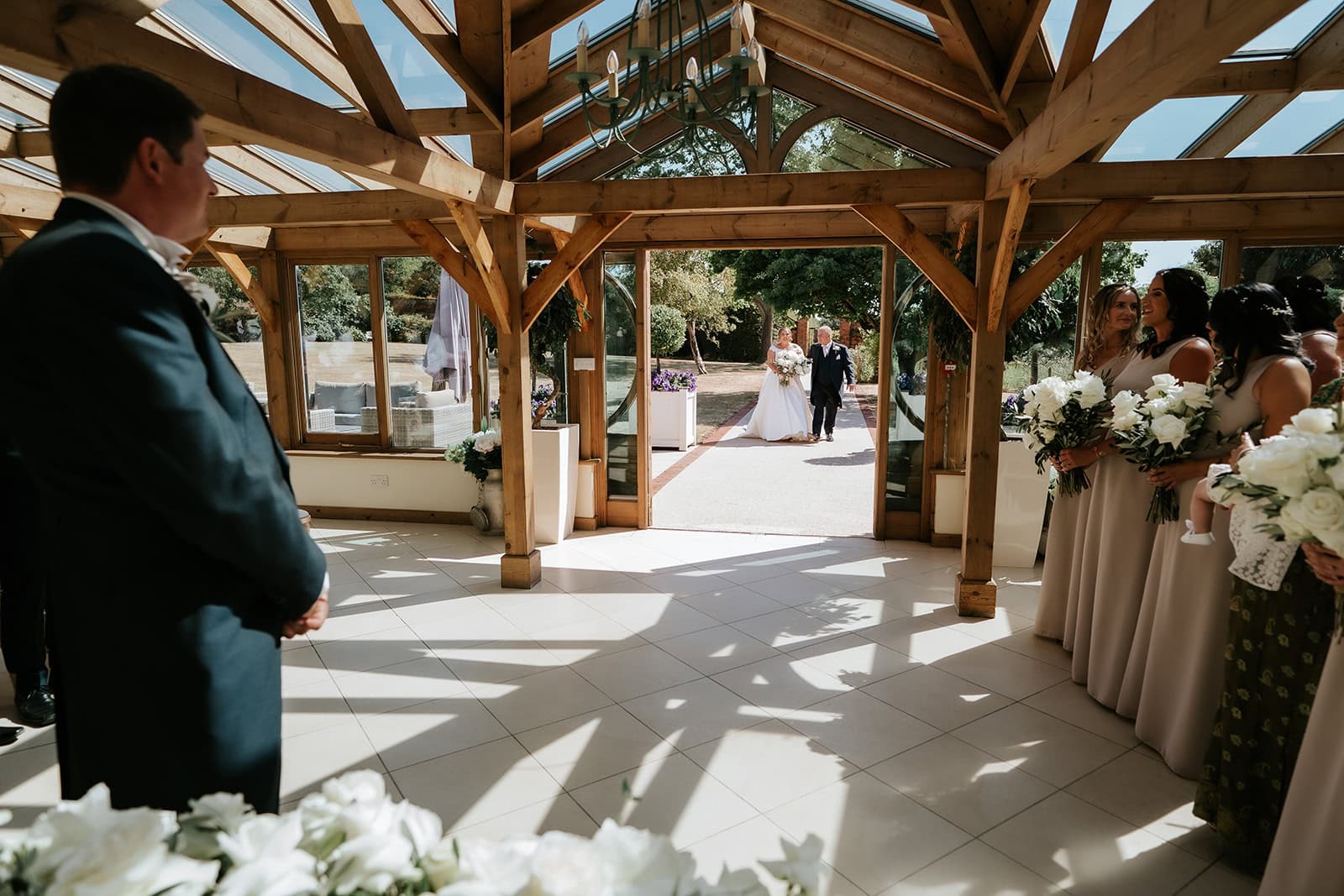 A groom waits at the altar inside the Orangery at Gaynes Park, Epping, Essex, as the bride appears at the entrance to the Long Walk aisle, framed by the oak doorway and garden beyond. Dramatic natural light falls across the tiled floor from the glass roof above. Photographed by Tel, Lily & White Photography, Gaynes Park wedding photographer.
