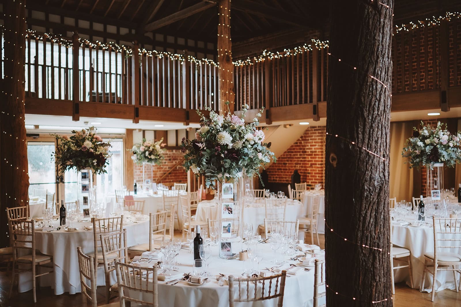 The Mill Barn reception room at Gaynes Park wedding venue in Epping, Essex, dressed for a wedding breakfast. Ancient oak trunks rise through the centre of the room beneath a mezzanine level strung with fairy lights, with tall floral centrepieces on round tables dressed in white linen. Photographed by Tel, Lily & White Photography, Gaynes Park wedding photographer.