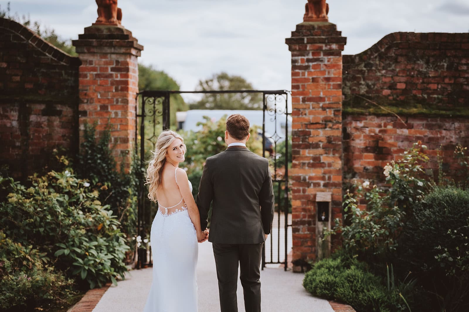 A bride looks back over her shoulder as she and her groom walk hand in hand toward the wrought iron gates of the Victorian Walled Garden along the Long Walk at Gaynes Park, Epping, Essex. The ancient brick gateposts and lush garden borders frame the couple from behind. Photographed by Tel, Lily & White Photography, Gaynes Park wedding photographer.
