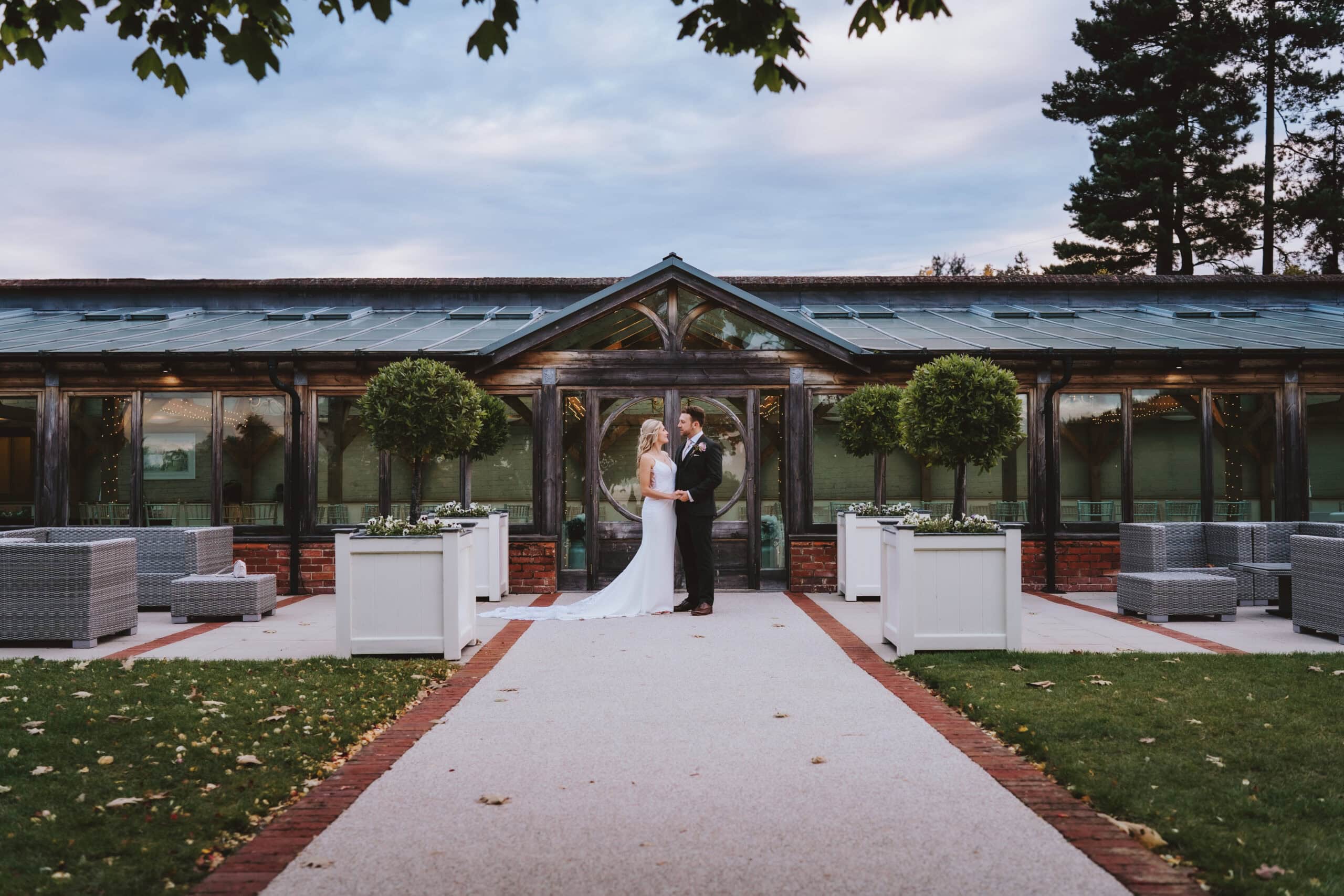 A bride and groom stand holding hands in front of the Orangery at Gaynes Park wedding venue in Epping, Essex. The couple are framed perfectly within the circular window of the Orangery entrance, surrounded by topiary trees and fairy lights. Photographed by Tel James, Lily & White Photography, Gaynes Park wedding photographer.
