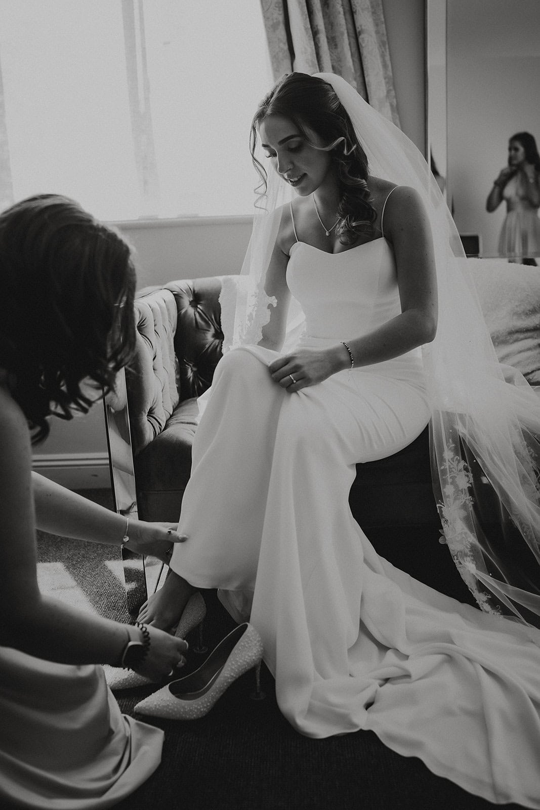 A cinematic black and white wedding photograph in Suffolk, capturing a quiet, editorial moment of a bride preparing for her ceremony at All Saints Hotel.