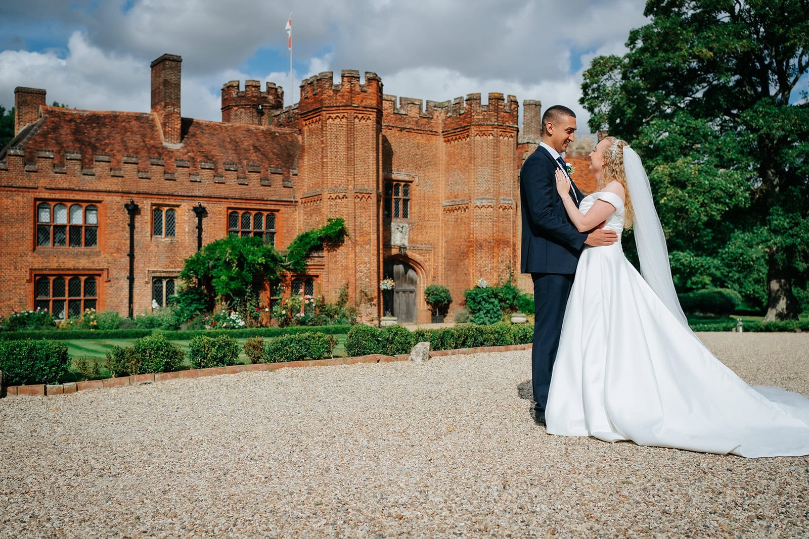 A bride and groom stand together on the gravel driveway in front of the full facade of Leez Priory, Little Leighs, Essex. The Grade I listed Tudor red brick manor house with its castellated tower rises behind the couple beneath a dramatic summer sky. Photographed by Tel, Lily & White Photography, Leez Priory wedding photographer.