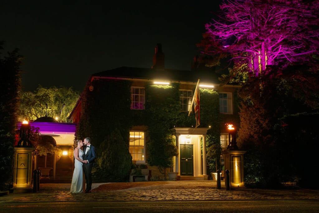 Bride and groom kissing outside the ivy-covered Friern Manor at night with purple up lighting and courtyard – evening wedding photography by Lily & White