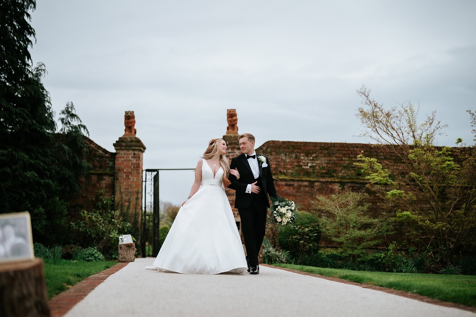 A bride and groom walk hand in hand along the Long Walk at Gaynes Park wedding venue in Epping, Essex. The ancient brick walls and stone gateposts of the Victorian Walled Garden frame the couple as they approach the Orangery. Photographed by Tel, Lily & White Photography, Gaynes Park wedding photographer.