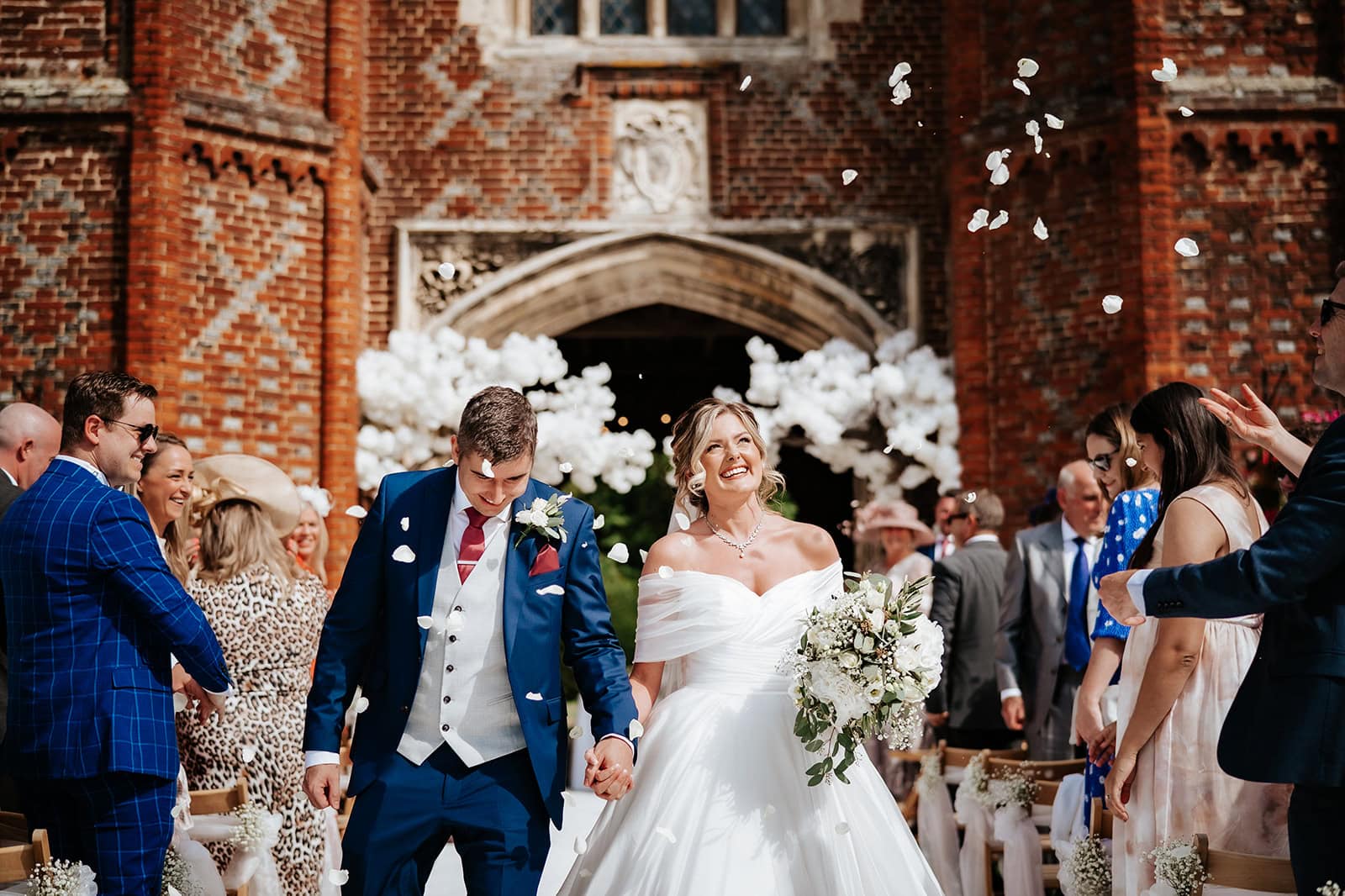 A bride and groom walk through a shower of white rose petal confetti in front of the Tudor carriageway arch at Leez Priory, Little Leighs, Essex. The bride laughs joyfully as guests line both sides of the path in bright summer sunshine. Photographed by Tel, Lily & White Photography, Leez Priory wedding photographer.