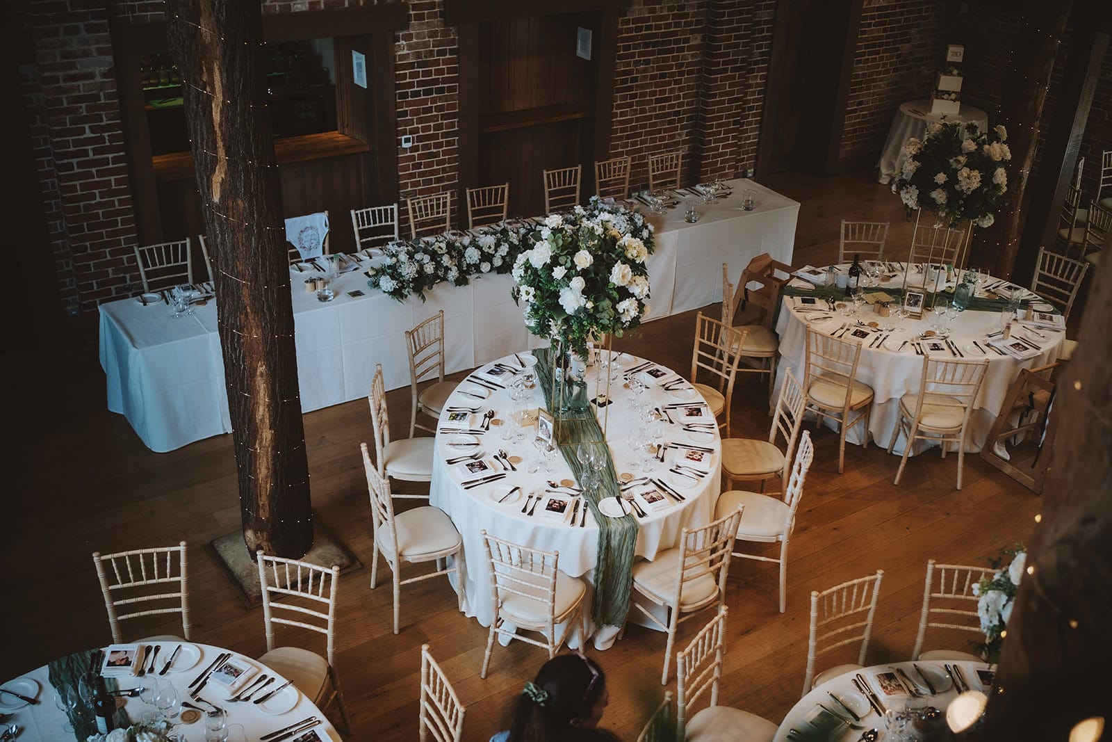 A top-down view from the mezzanine level of the Mill Barn at Gaynes Park, Epping, Essex, showing the wedding breakfast layout with a traditional top table, round guest tables and tall floral arrangements. The ancient oak trunks and exposed brick walls of the barn are visible throughout. Photographed by Tel, Lily & White Photography, Gaynes Park wedding photographer.