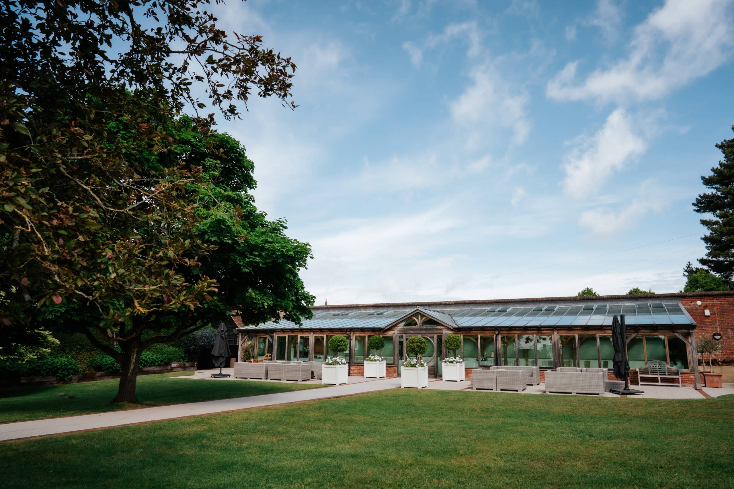 The exterior of the Orangery at Gaynes Park wedding venue in Epping, Essex. The glass and timber structure sits within the Victorian Walled Garden, surrounded by manicured lawns, mature trees and white floral planters. Photographed by Tel, Lily & White Photography, Gaynes Park wedding photographer.