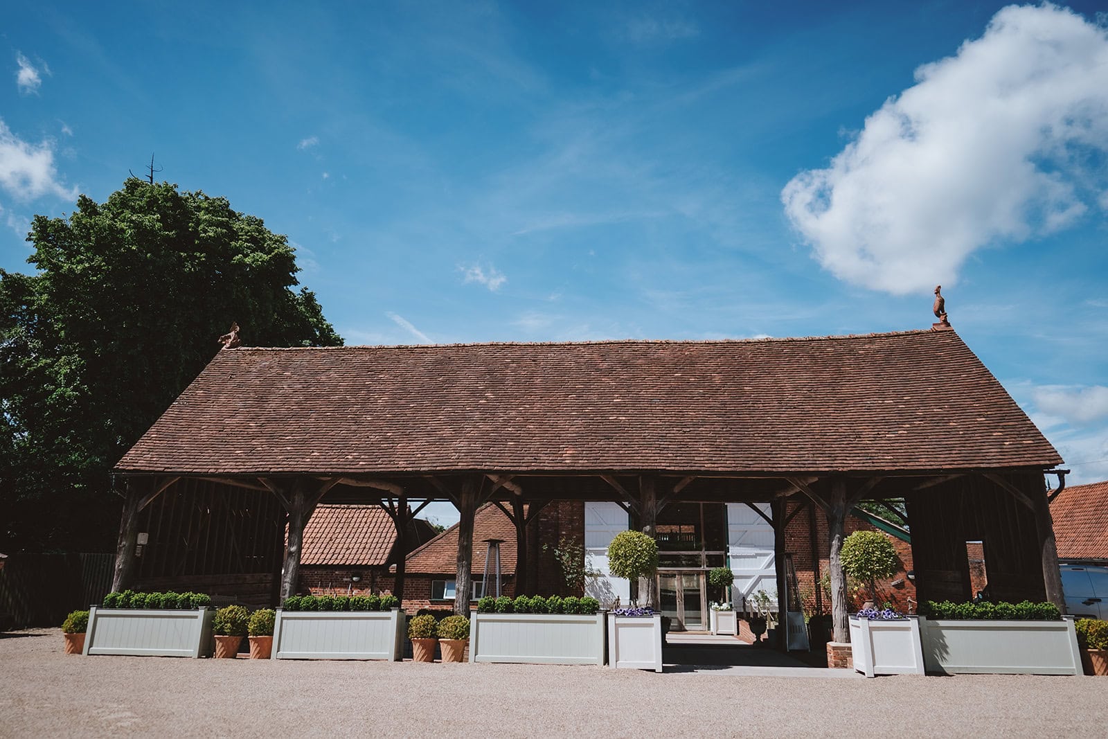 The exterior of the Gather Barn at Gaynes Park wedding venue in Epping, Essex. The open-sided timber and brick structure sits beneath a traditional clay tile roof on a sunny summer day, flanked by box hedging, topiary trees and sage green planters. Photographed by Tel, Lily & White Photography, Gaynes Park wedding photographer.