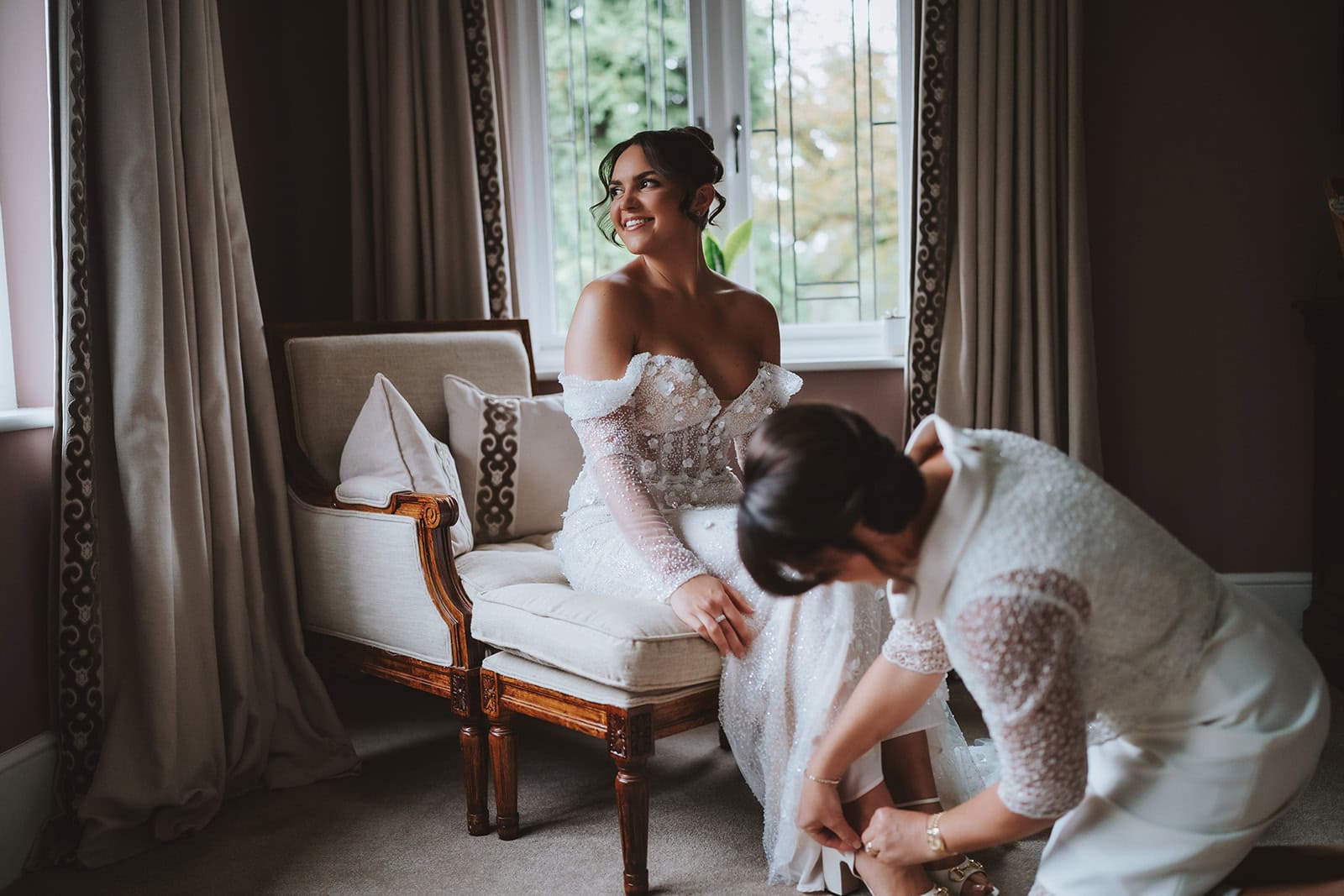 Abbey seated by the window in her off-shoulder beaded wedding gown while having her shoes fastened during bridal preparations at Blake Hall – wedding photography by Tel, Lily & White