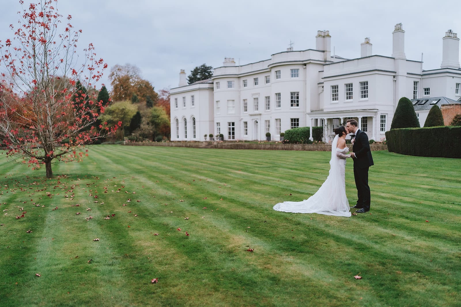 Abbey and Charlie kissing on the lawn in front of the white Georgian manor house at Blake Hall, autumn foliage in the background – Blake Hall wedding photography by Tel, Lily & White