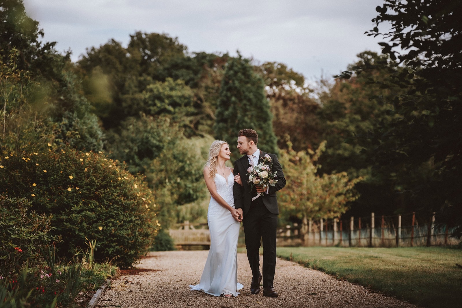 Bride and groom walking hand in hand along a tree-lined gravel path in the gardens at Gaynes Park, Essex, the groom carrying her bouquet as they share a glance — wedding photography by Lily & White