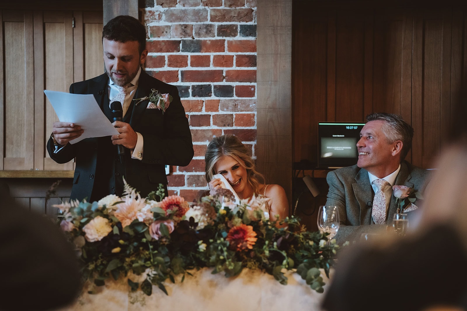 Harvey delivering his wedding speech in the Mill Barn at Gaynes Park, Essex, as Emily wipes away tears of laughter at the top table – 29th September 2024, photographed by Tel, Lily & White