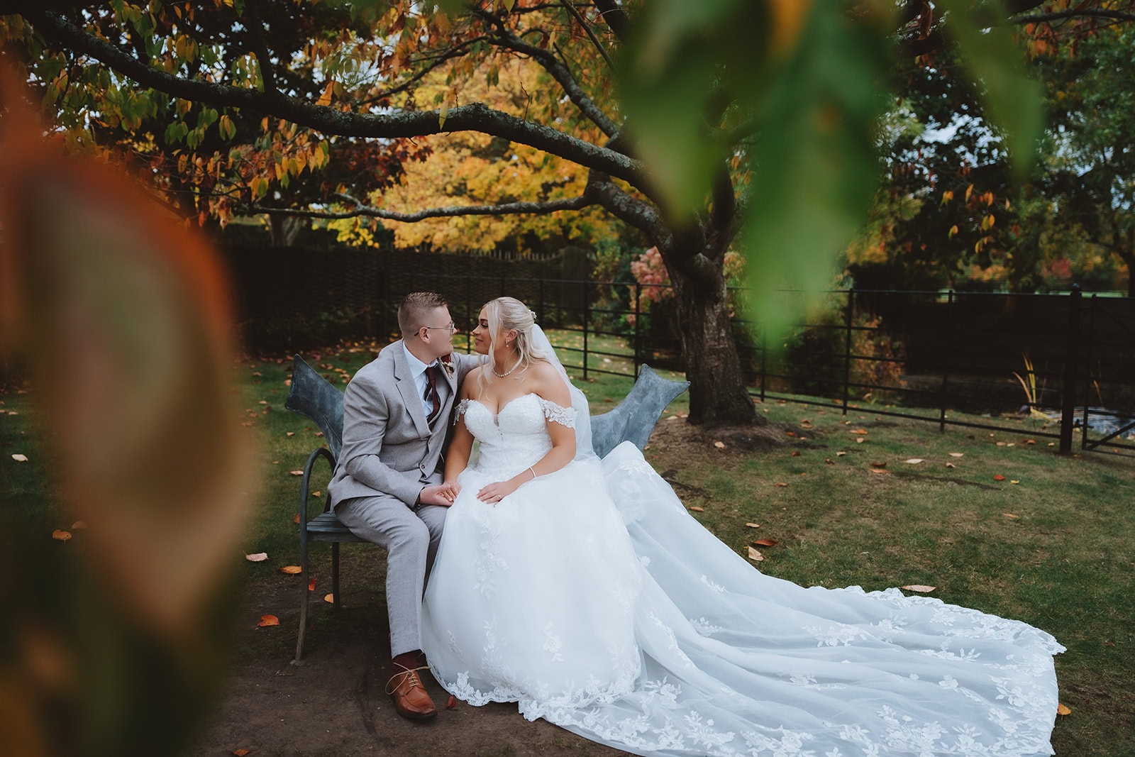 Holly and Billy sitting together on a bench in the grounds of Blake Hall, framed by autumn foliage in golden and russet tones, October 2025 – wedding photography by Tel, Lily & White