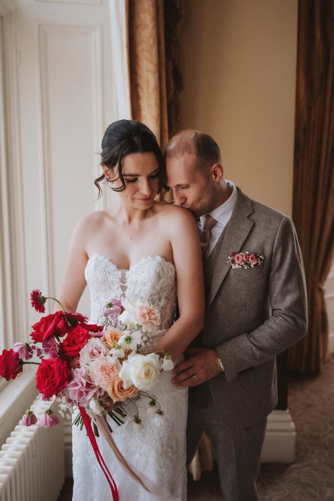 Editorial wedding photography Essex featuring intimate couple portrait with bride in strapless lace dress and groom in tweed suit with window light