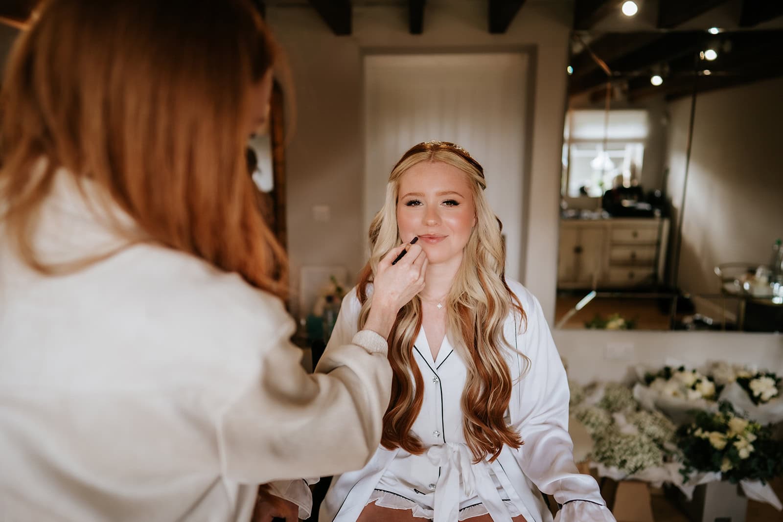 Olivia smiling as her makeup is applied during bridal preparations at Gaynes Park, wearing a white bridal robe, floral arrangements visible in the background – wedding photography by Tel, Lily & White