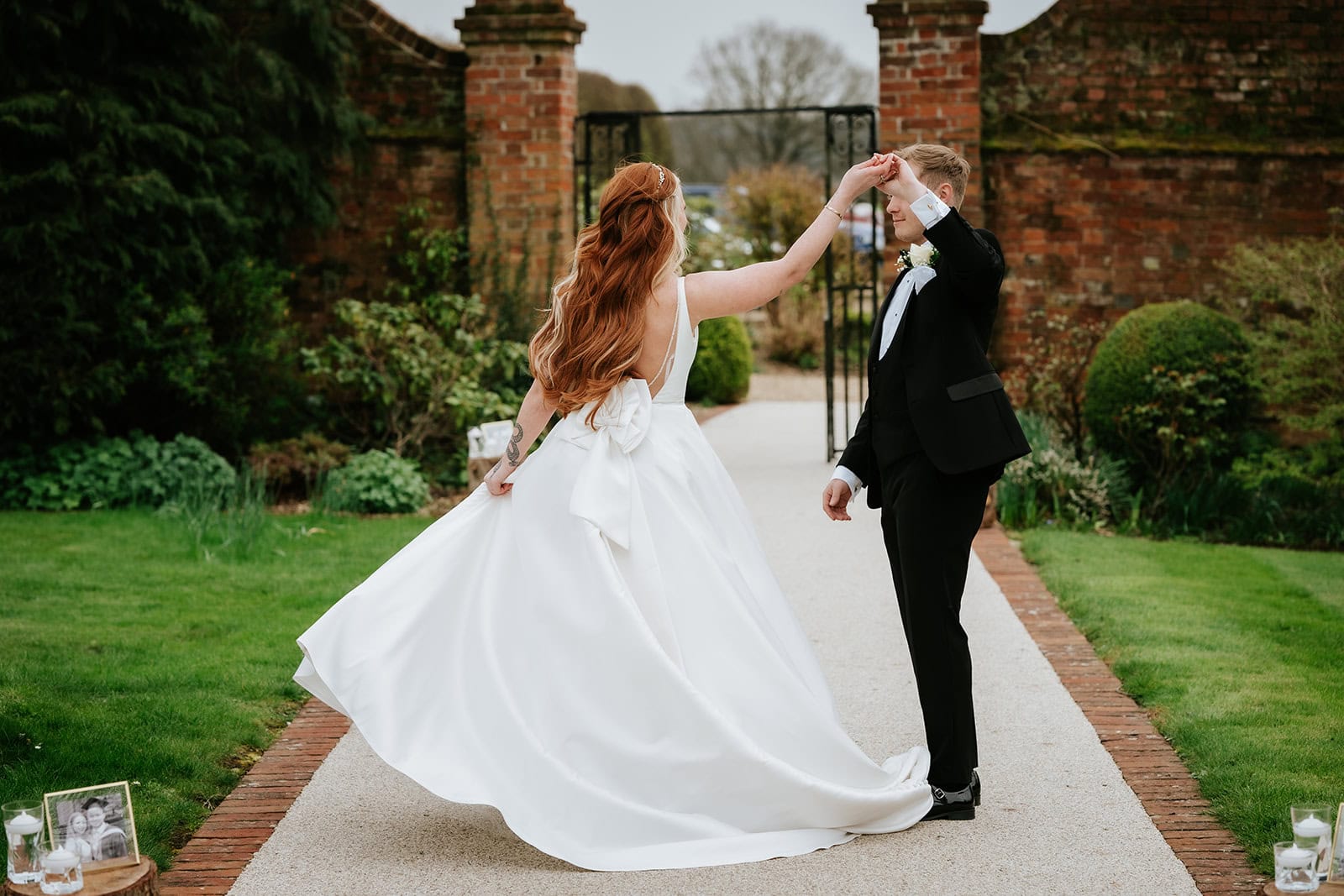 Olivia being spun by Alfie on the path through the walled garden at Gaynes Park, her ballgown skirt sweeping wide, framed photographs and candles lining the path – wedding photography by Tel, Lily & White
