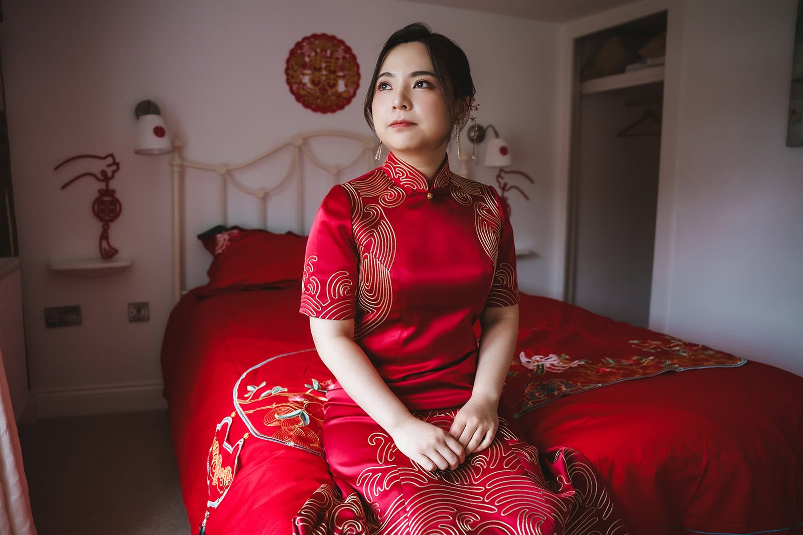 Yiran seated on the bed in her red embroidered qipao during bridal preparations at Blake Hall, traditional Chinese double happiness decorations on the wall behind her – wedding photography by Tel, Lily & White