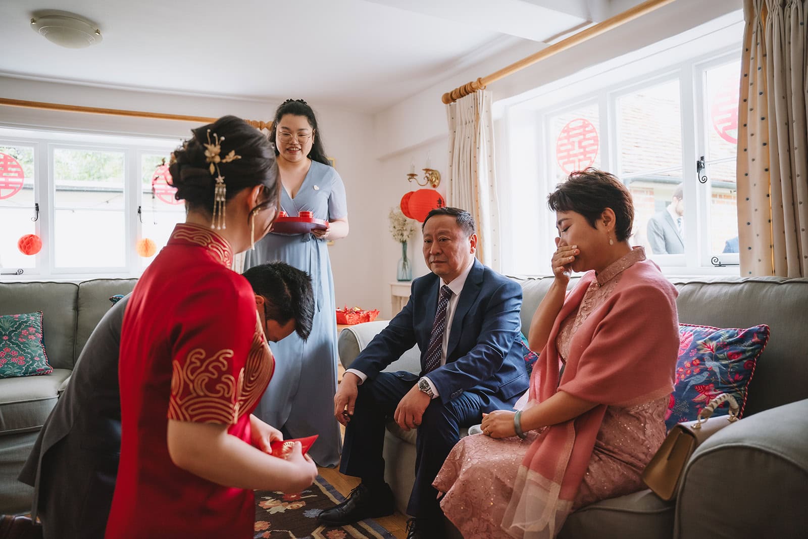 Yiran and Jeffrey presenting tea to family during the Chinese tea ceremony at Blake Hall, Yiran's mother visibly emotional, red lanterns and double happiness decorations in the background – wedding photography by Tel, Lily & White