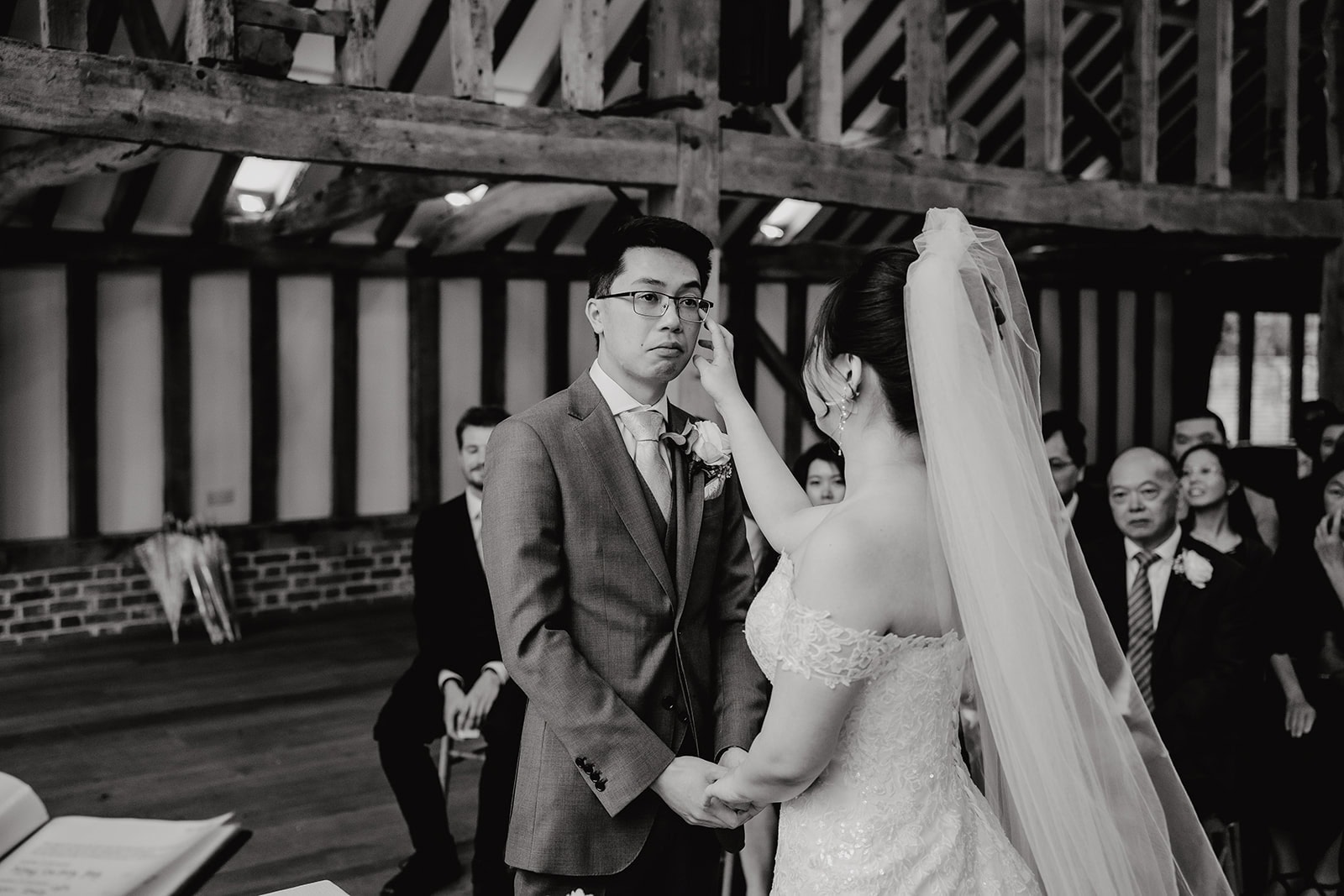 Yiran wiping tears from Jeffrey's eyes during their wedding ceremony in the Hay Barn at Blake Hall, guests smiling in the background – wedding photography by Tel, Lily & White